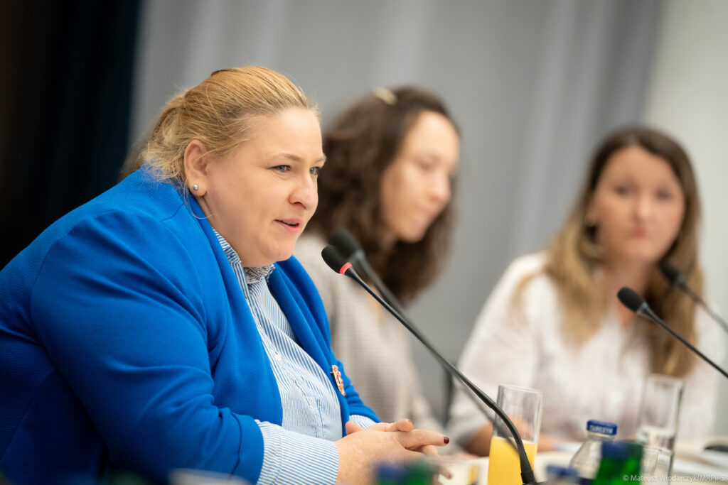 Wiceministra Maja Nowak podczas spotkania konsultacyjnego dotyczącego zatrudnienia wspomaganego osób z niepełnosprawnościami. Siedzi przy stole konferencyjnym i rozmawia z uczestnikami spotkania.