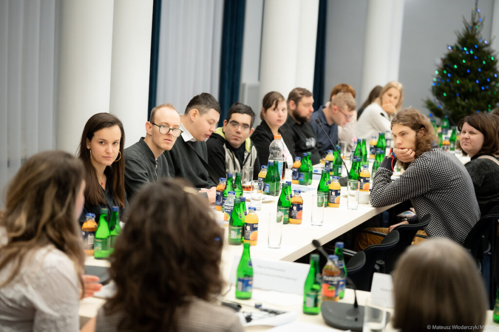 Uczestnicy spotkania konsultacyjnego na temat zatrudnienia wspomaganego osób z niepełnosprawnościami, siedzący przy stołach w sali konferencyjnej i biorący udział w dyskusji.