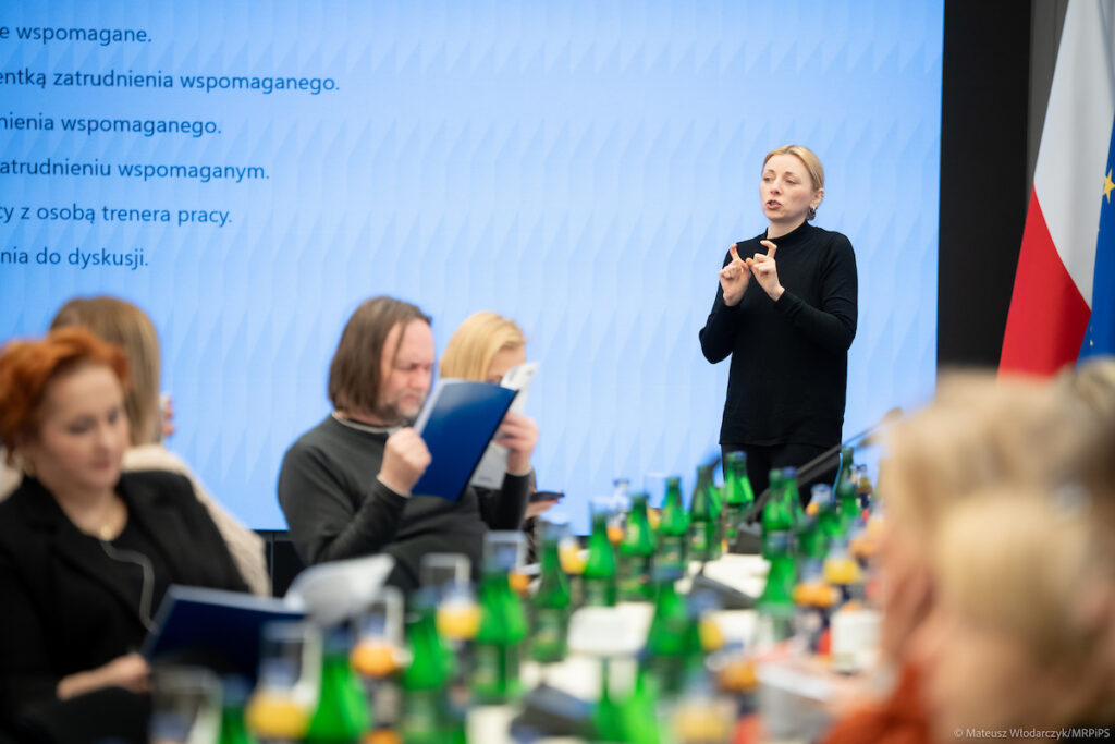 Tłumacz języka migowego podczas spotkania konsultacyjnego dotyczącego zatrudnienia wspomaganego, widoczny na pierwszym planie; w tle uczestnicy spotkania biorący udział w dyskusj