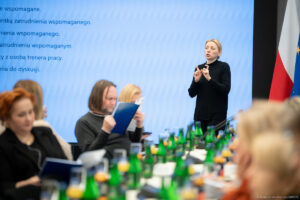 Tłumacz języka migowego podczas spotkania konsultacyjnego dotyczącego zatrudnienia wspomaganego, widoczny na pierwszym planie; w tle uczestnicy spotkania biorący udział w dyskusj