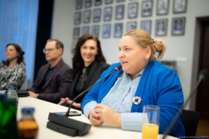 Wiceministra Maja Nowak podczas spotkania konsultacyjnego dotyczącego zatrudnienia wspomaganego osób z niepełnosprawnościami. Siedzi przy stole konferencyjnym i rozmawia z uczestnikami spotkania.