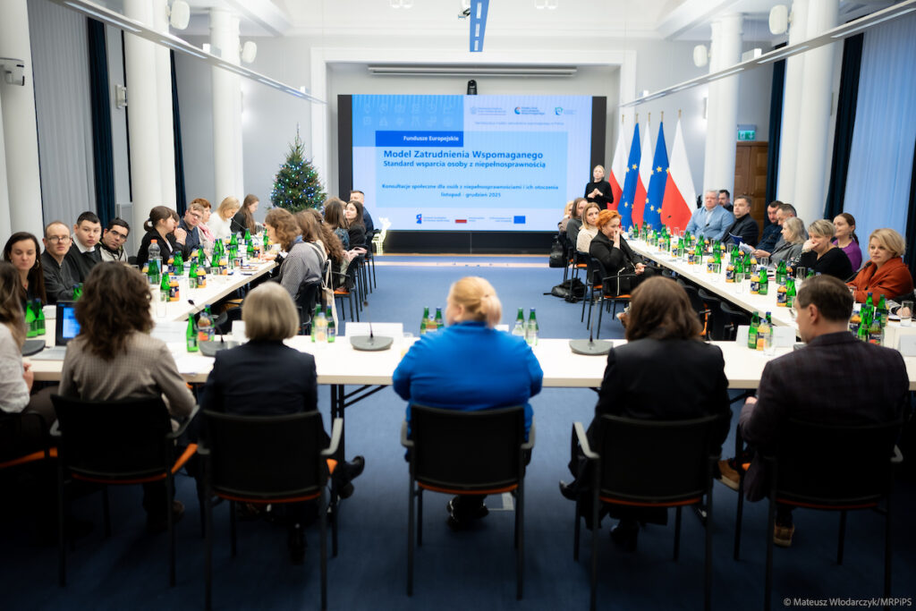 Uczestnicy spotkania konsultacyjnego na temat zatrudnienia wspomaganego osób z niepełnosprawnościami, siedzący przy stołach w sali konferencyjnej i biorący udział w dyskusji.