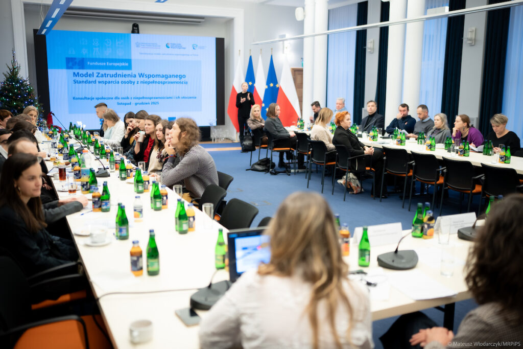 Uczestnicy spotkania konsultacyjnego na temat zatrudnienia wspomaganego osób z niepełnosprawnościami, siedzący przy stołach w sali konferencyjnej i biorący udział w dyskusji.