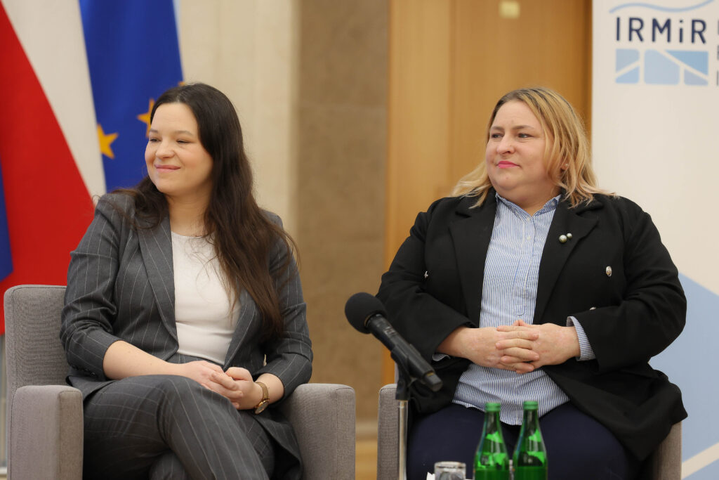 Na zdjęciu widać wiceministry pracy: Maję Nowak oraz Katarzynę Nowakowską, które uczestniczą w konferencji. W tle flagi polskie i unijne.