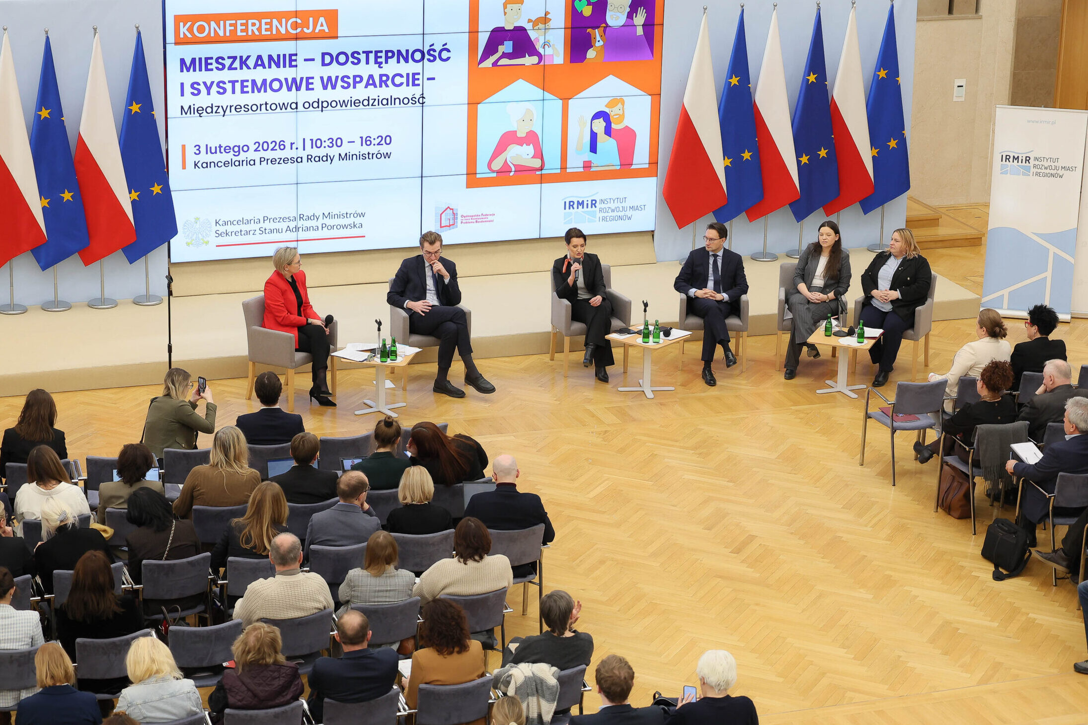 Na zdjęciu widać sześciu uczestników panelu dyskusyjnego siedzących przed liczną widownią. W tle flagi polskie i unijne oraz ekran na którym wyświetlona jest nazwa konferencji: Mieszkalnictwo - dostępność i systemowe wsparcie