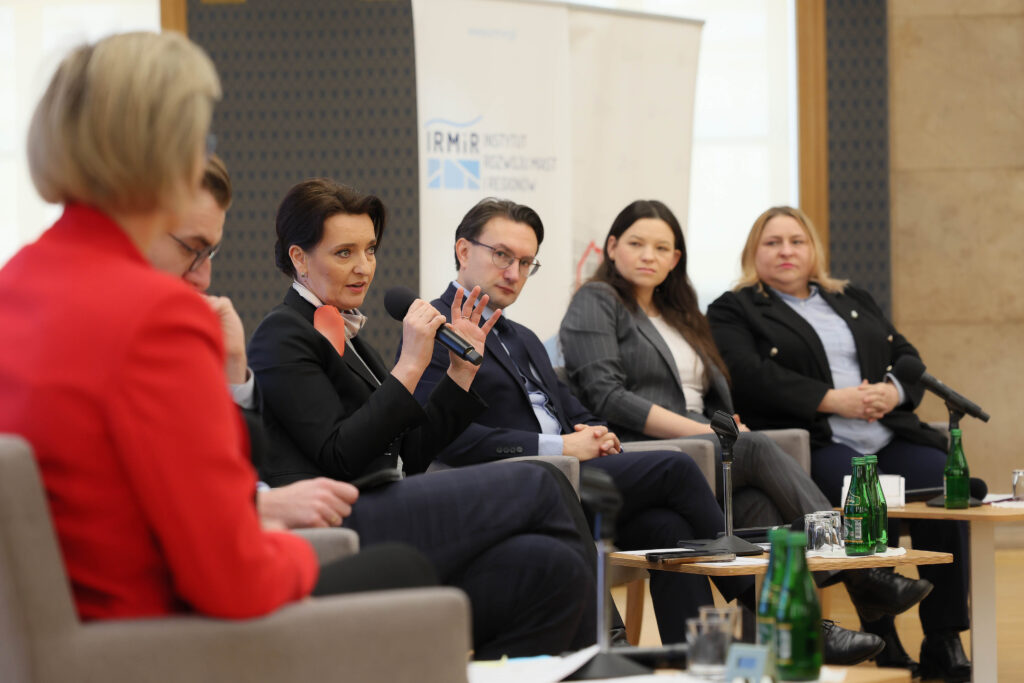 Na zdjęciu widać uczestników panelu dyskusyjnego siedzących przy stolikach. W tle flagi polskie i unijne oraz ekran na którym wyświetla się plakat konferencji.