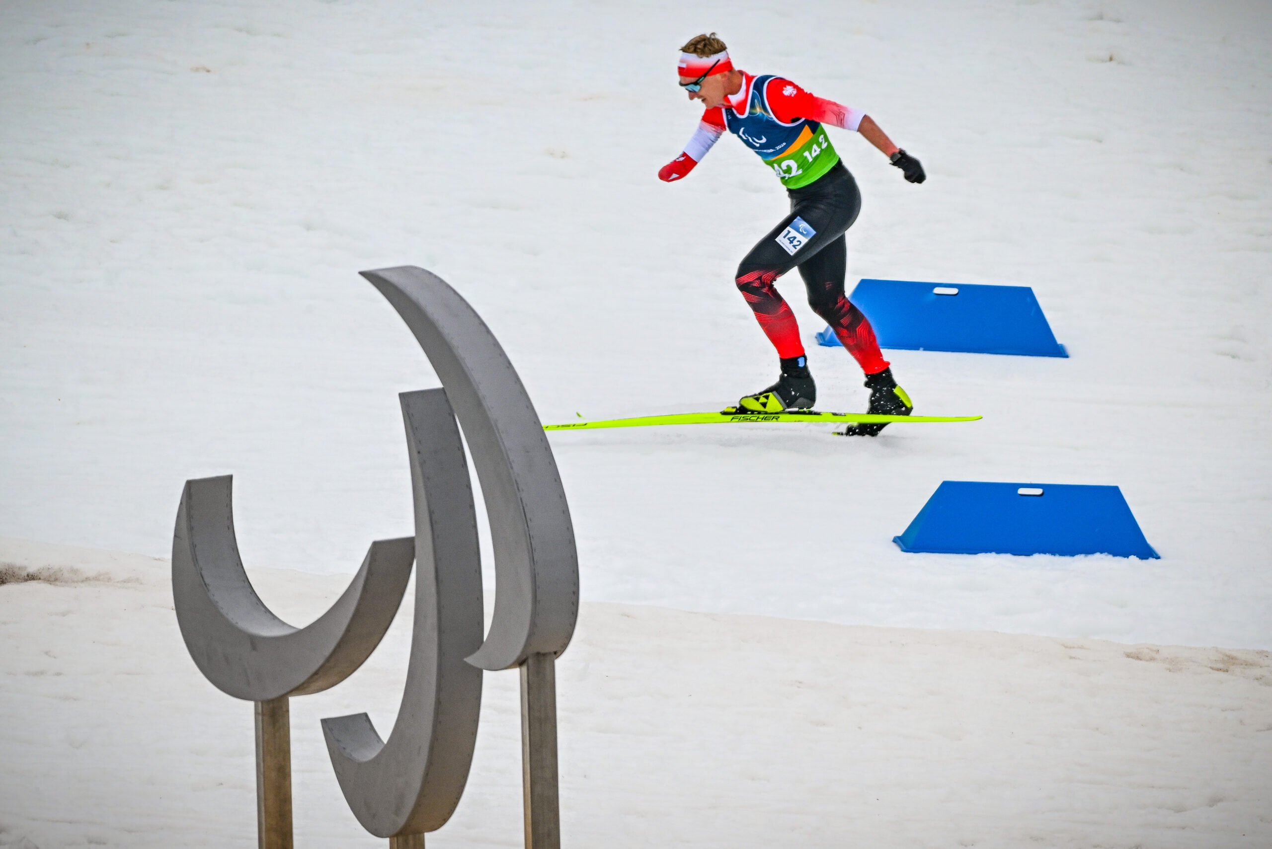 Reprezentant Polski, Witold Skupień, w podbiegu na wzniesienie podczas biegu narciarskiego. Witold biegnie bez użycia kijów narciarskich (nie ma obydwu dłoni). Na pierwszym planie dekoracyjnie wkomponowany w kadr zrobiony ze stali znak igrzysk paralimpijskich, tzw. Agitos.