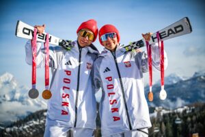 dwóch reprezentantów Polski w narciarstwie alpejskim, Michał Gołaś i Kacper Walas stoi w strojach reprezentacyjnych stoi na tle gór, za plecami trzymają narty, a na nich w zawadiacki sposób zawieszono zdobyte przez nich srebrne i brązowe medale.