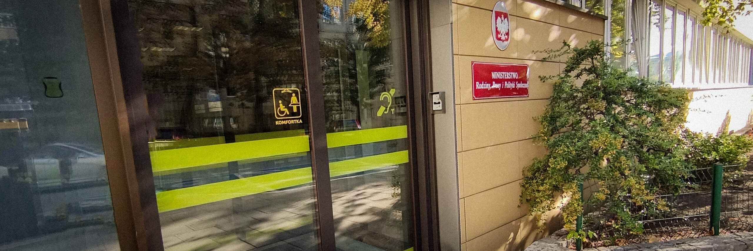 Biuro Pełnomocnika 1 Zdjęcie przedstawia wejście do siedziby Biura. Na drzwiach znajduje się informacja o dostępnej komfortce oraz pętli indukcyjnej. Obok drzwi tablica z napisem Ministerstwo Rodziny, Pracy i Polityki Społecznej