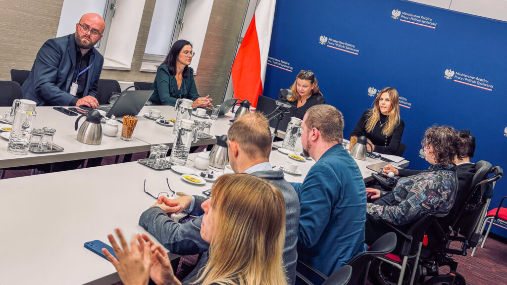 Uczestnicy posiedzenia Rady Programowej Centrum Komunikacji podczas obrad przy wspólnym stole. Jedna z uczestniczek porusza się na wózku. W tle ścianka z logotypami MRPiPS.