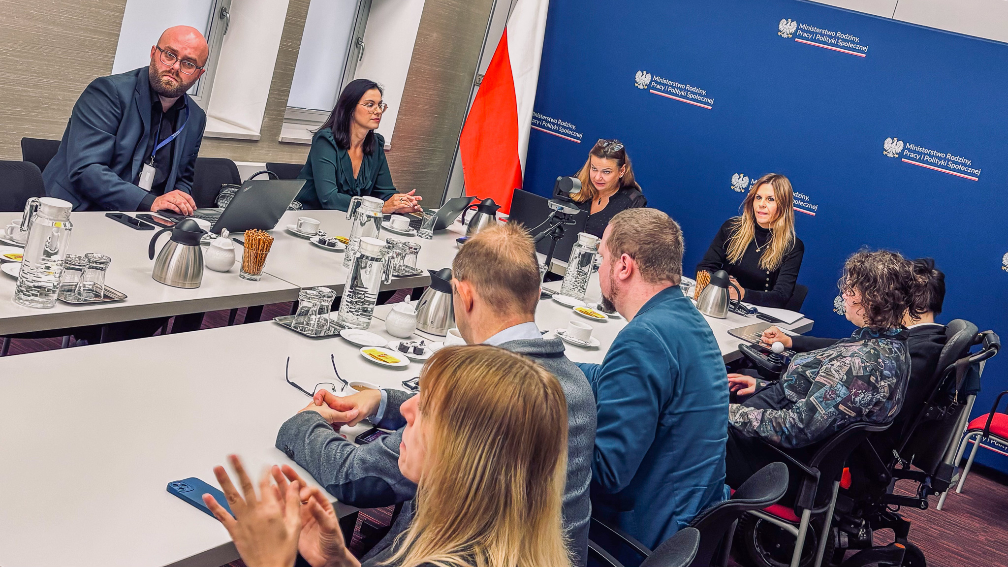 Uczestnicy posiedzenia Rady Programowej Centrum Komunikacji podczas obrad przy wspólnym stole. Jedna z uczestniczek porusza się na wózku. W tle ścianka z logotypami MRPiPS.