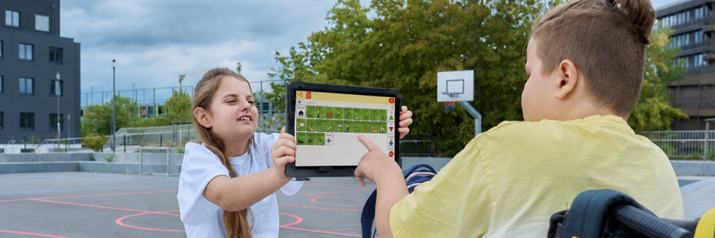 Na boisku sportowym dziewczynka w białej koszulce trzyma tablet z tablicą komunikacyjną zawierającą symbole graficzne. Chłopiec w żółtej koszulce, siedzący na wózku, wskazuje palcem jeden z symboli na ekranie. W tle widoczne są drzewa, kosz do koszykówki i budynki.