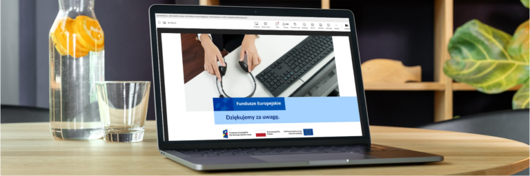 Na drewnianym stole stoi otwarty laptop, na którego ekranie widać końcowy slajd prezentacji dotyczącej zatrudnienia wspomaganego. Widoczny jest napis „Dziękujemy za uwagę” oraz logotypy Funduszy Europejskich, Rzeczypospolitej Polskiej i Unii Europejskiej. Obok laptopa stoją szklanka i karafka z wodą oraz plasterkami pomarańczy.