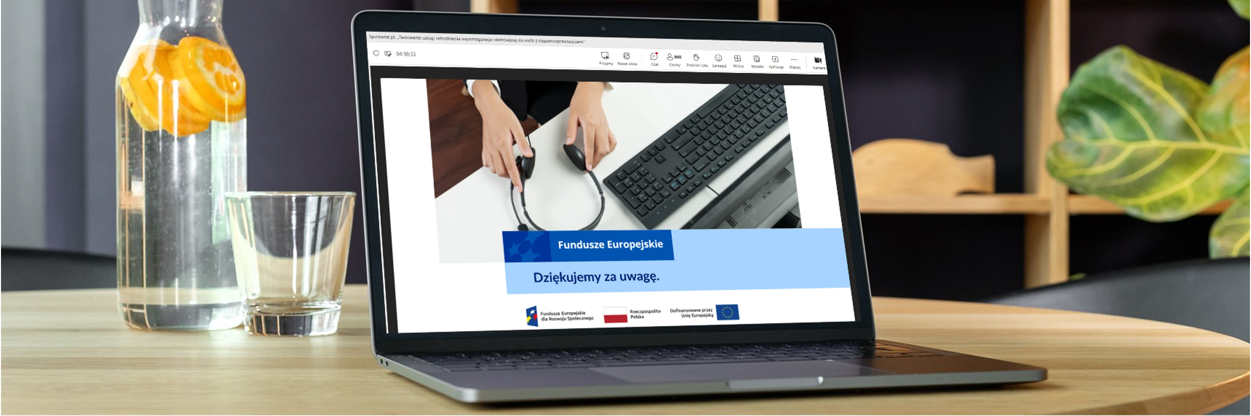 Na drewnianym stole stoi otwarty laptop, na którego ekranie widać końcowy slajd prezentacji dotyczącej zatrudnienia wspomaganego. Widoczny jest napis „Dziękujemy za uwagę” oraz logotypy Funduszy Europejskich, Rzeczypospolitej Polskiej i Unii Europejskiej. Obok laptopa stoją szklanka i karafka z wodą oraz plasterkami pomarańczy.