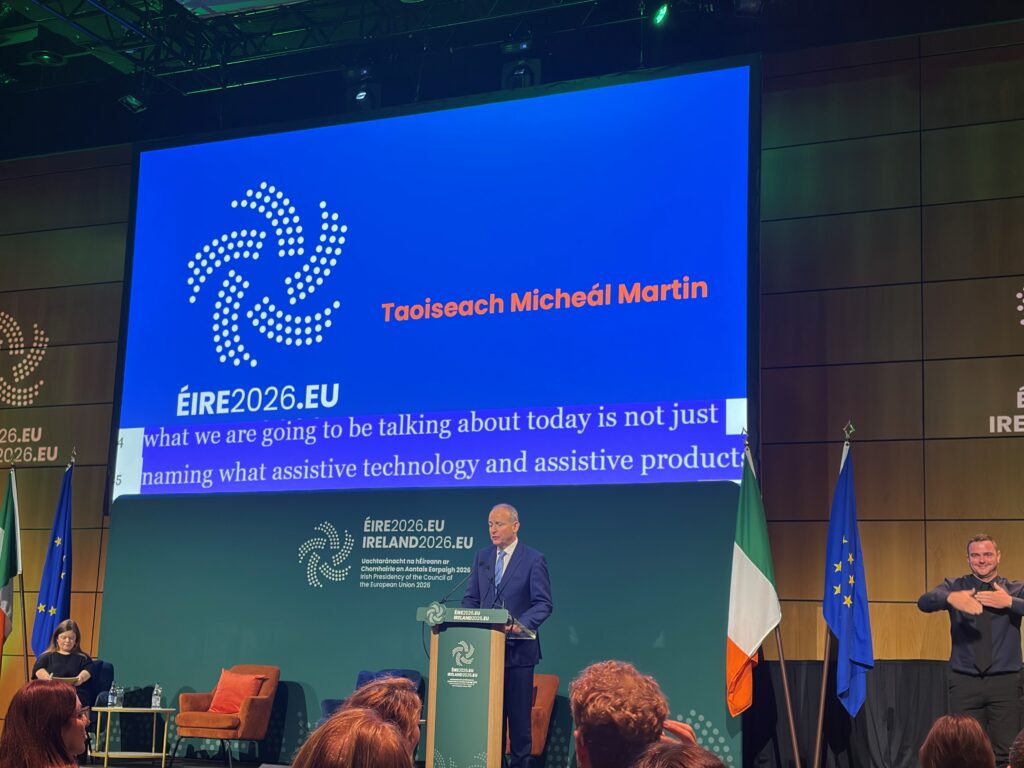 Premier Irlandii Micheál Martin przemawia przy mównicy podczas konferencji w Dublinie. Obok sceny stoi tłumacz języka migowego. W tle widoczne są flagi Irlandii i Unii Europejskiej oraz ekran z identyfikacją wizualną irlandzkiej prezydencji w Radzie UE.