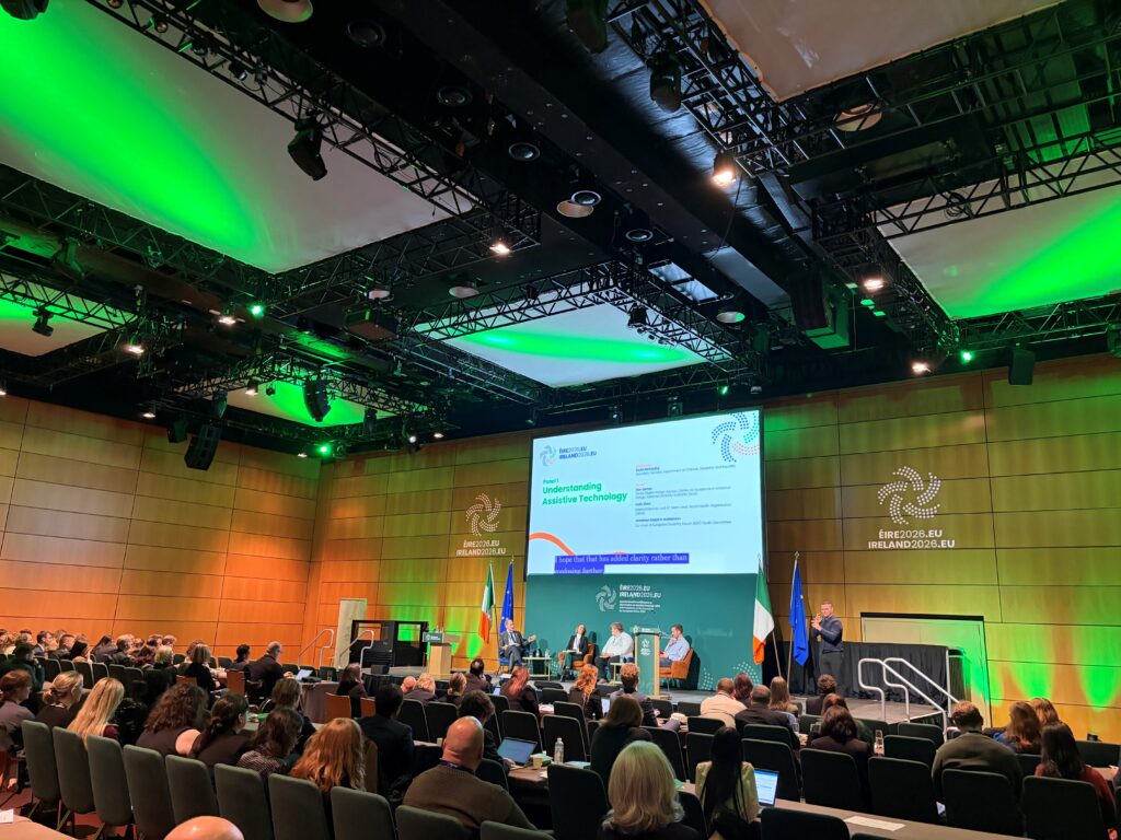 Widok na salę konferencyjną w Dublinie. Na scenie trwa dyskusja panelowa, którą obserwują uczestnicy wydarzenia. Sala jest oświetlona zielonym światłem, a na ekranie wyświetlany jest program konferencji.