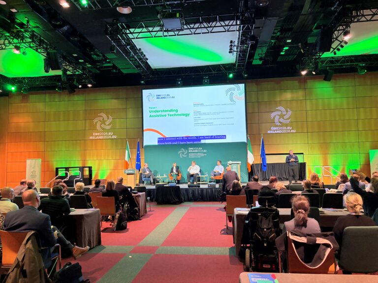 Uczestnicy konferencji w Dublinie obserwują dyskusję panelową poświęconą technologiom wspomagającym. Na scenie siedzą paneliści, a na dużym ekranie wyświetlany jest tytuł sesji „Understanding Assistive Technology”. Po prawej stronie sceny widoczny jest tłumacz języka migowego.